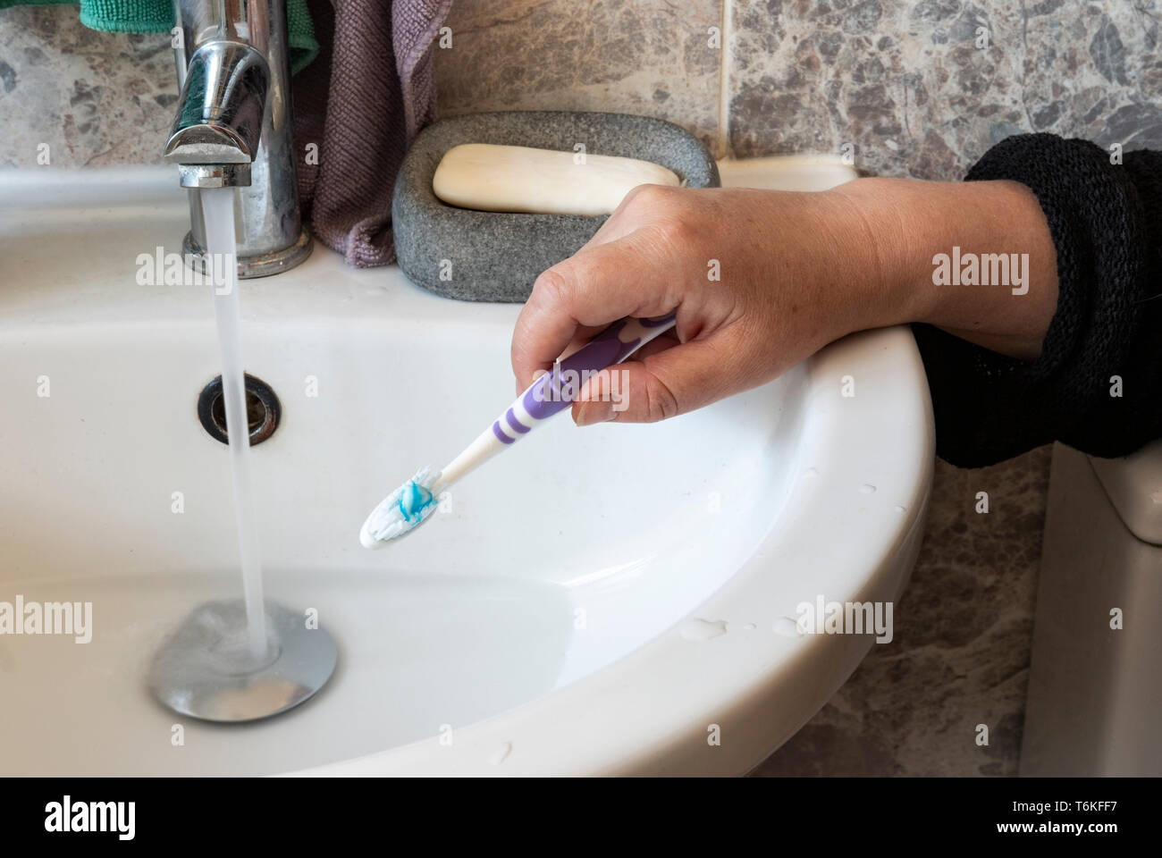 Hand, eine Zahnbürste und Zahnpasta auf, neben dem Waschbecken, mit der Tap laufen gelassen. Stockfoto