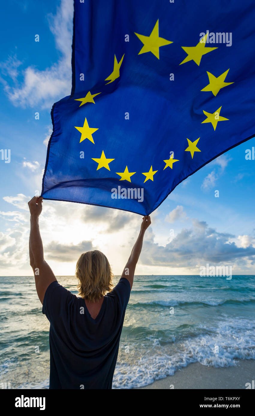 Mann mit blonden Haaren holding EU Europäische Union Flagge im Wind auf einem Sonnenaufgang Strand am Mittelmeer Stockfoto