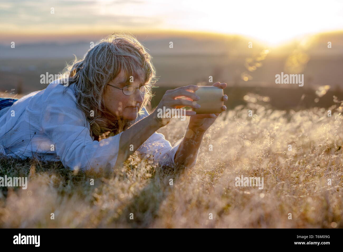 Liegende Frau in trockenem Gras und unter (selfie) Bilder mit dem Smartphone in der Natur bei Sonnenuntergang Stockfoto