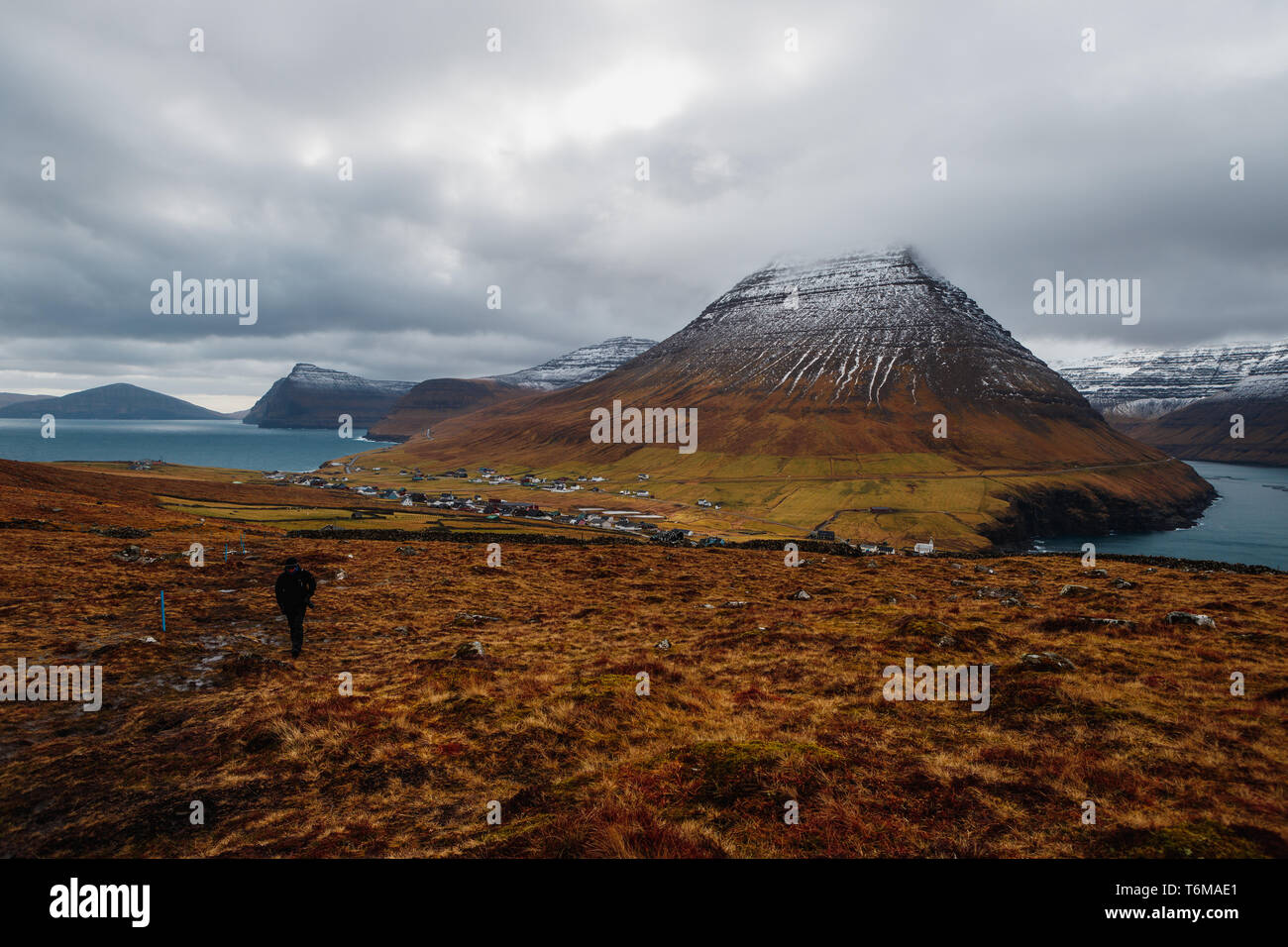 Einsamer Wanderer auf dem Weg an die Spitze von Kap Enniberg mit Blick auf Viðareiði und seine Fjord mit schneebedeckten Bergen (Färöer, Dänemark, Europa) Stockfoto