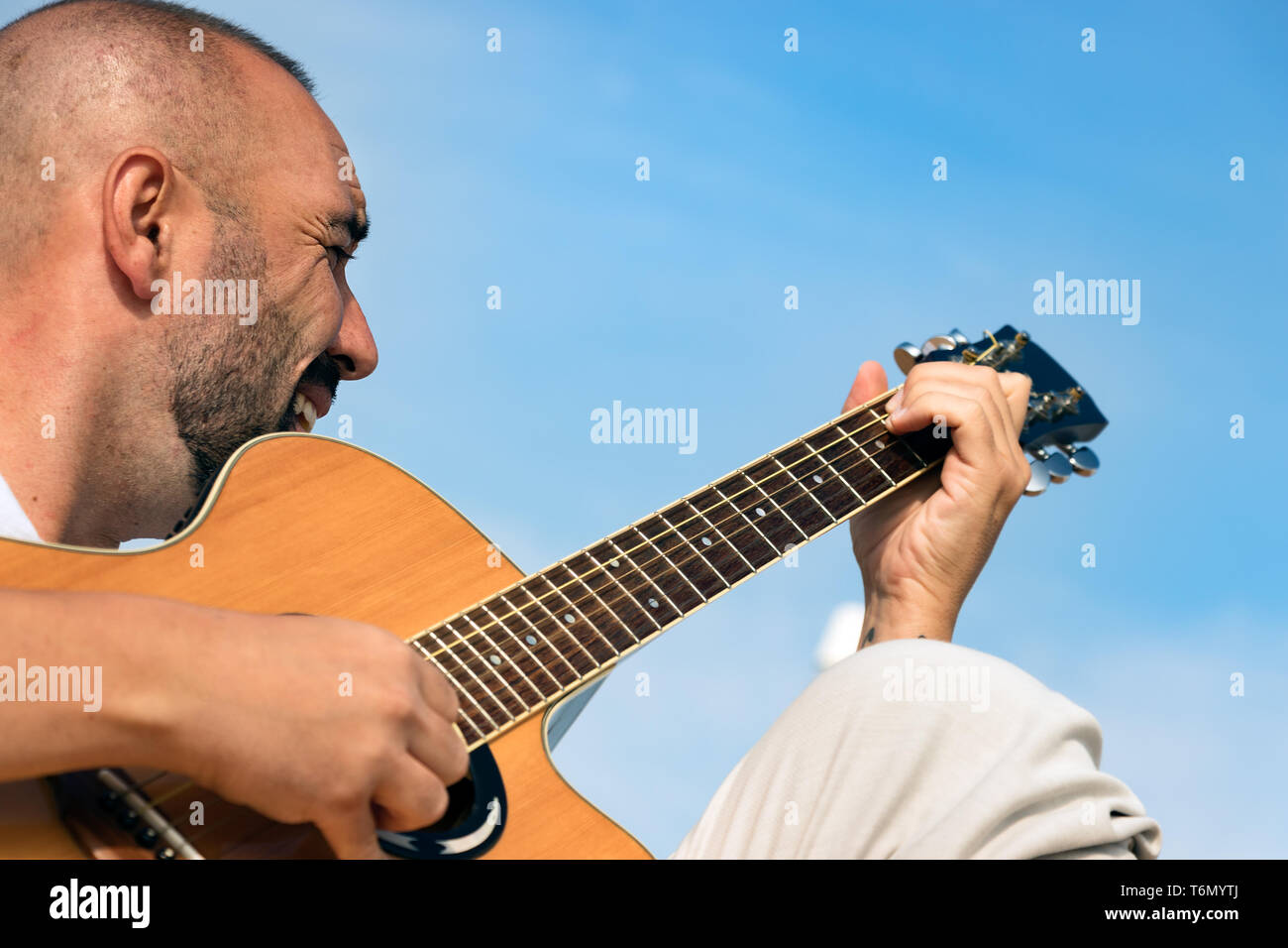 Porträt eines spanischen Mann mit Bart und kurzen buzz Haar, eine akustische Gitarre spielen im Freien über einen blauen Himmel Hintergrund. Stockfoto