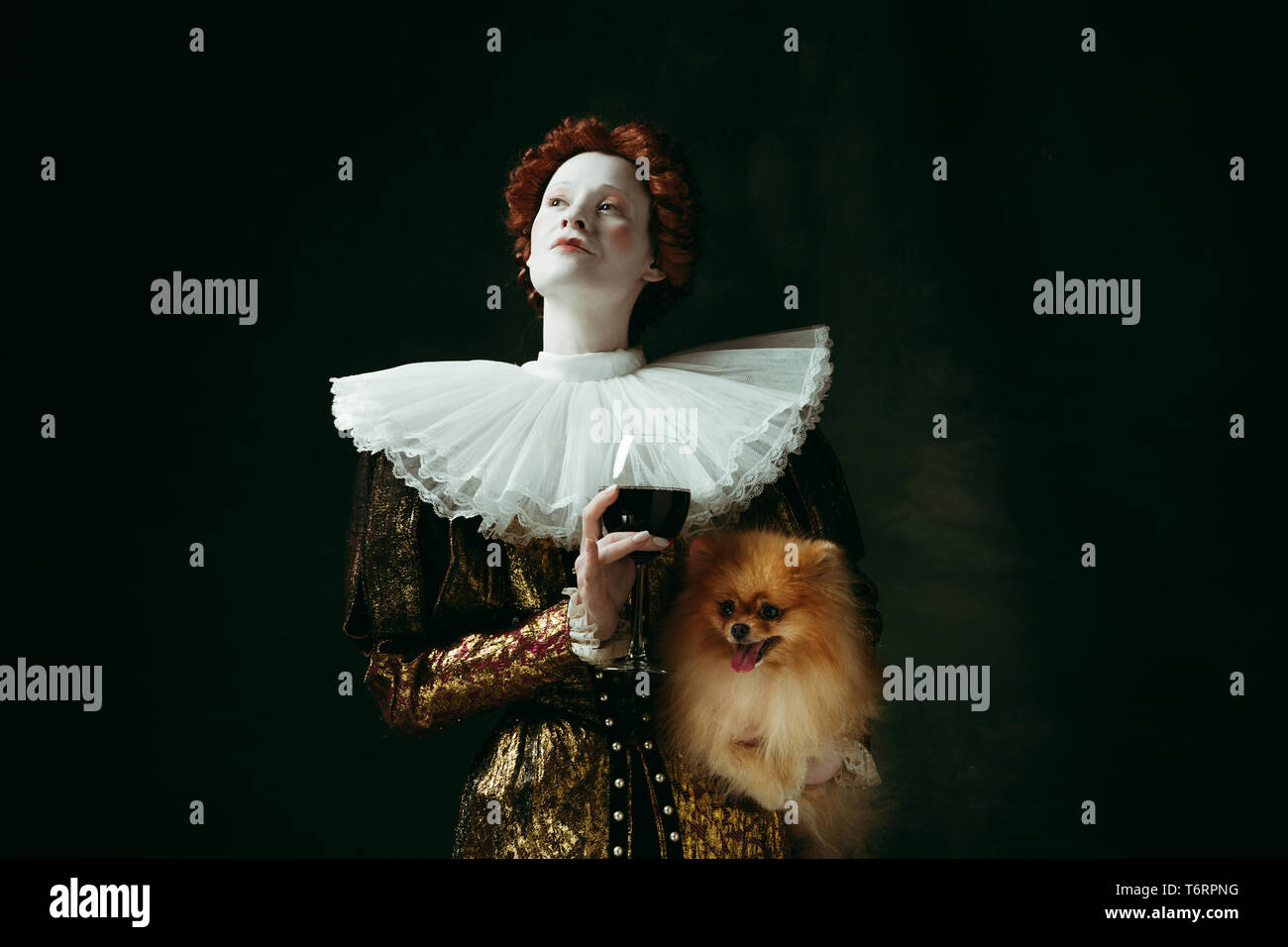 Werden sie sich wohlfühlen. Mittelalterliche rothaarige junge Frau im goldenen Vintage Kleidung als Herzogin holding Welpen und Glas mit Rotwein auf dunkelgrünem Hintergrund. Konzept der Vergleich der Epochen, Moderne, Renaissance. Stockfoto