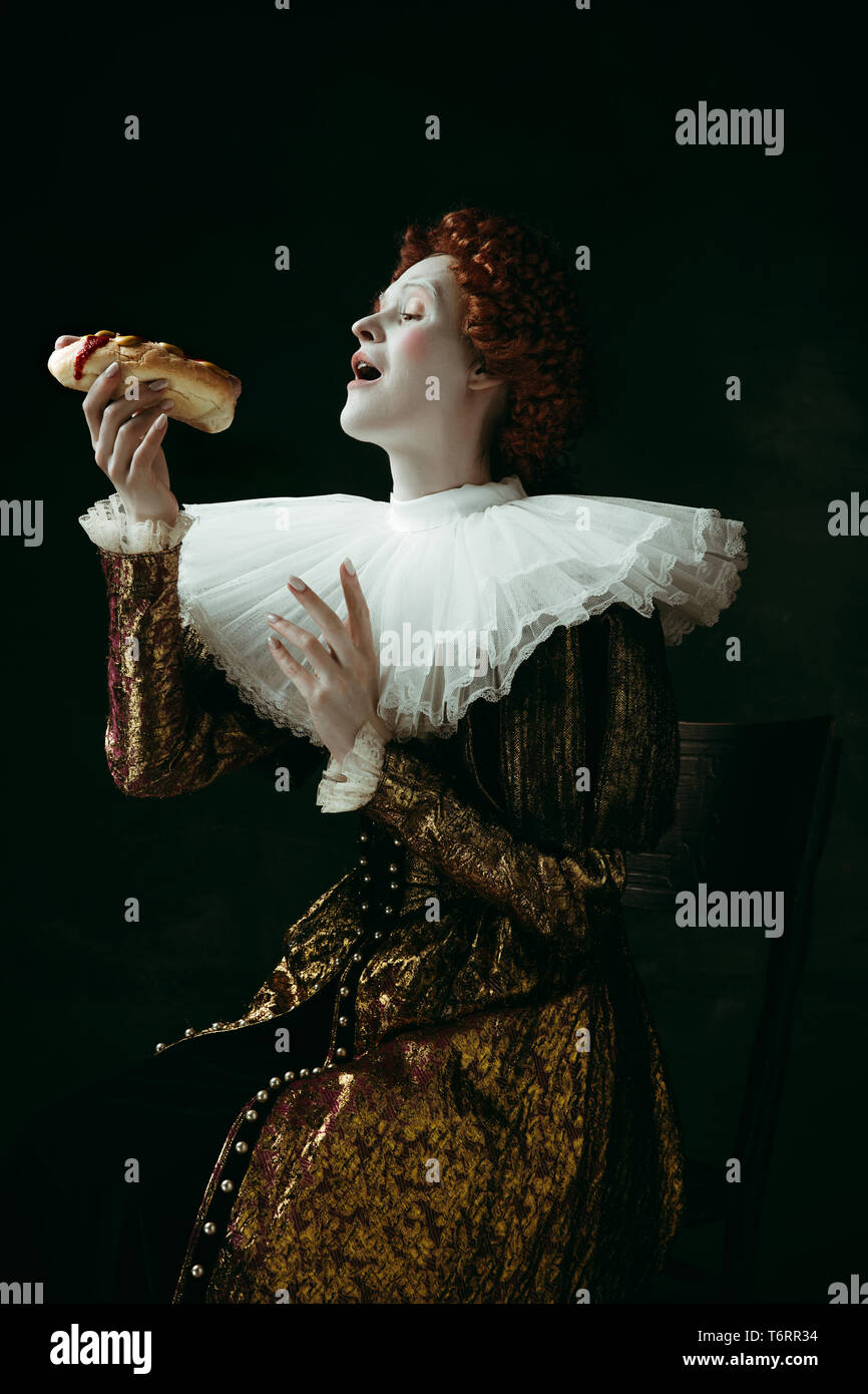 Ein kleiner Trick. Mittelalterliche rothaarige junge Frau im goldenen Vintage Kleidung als Herzogin essen einen Hot dog oder Sandwich auf dunkelgrünem Hintergrund. Konzept der Vergleich der Epochen, Modernität und Renaissance. Stockfoto