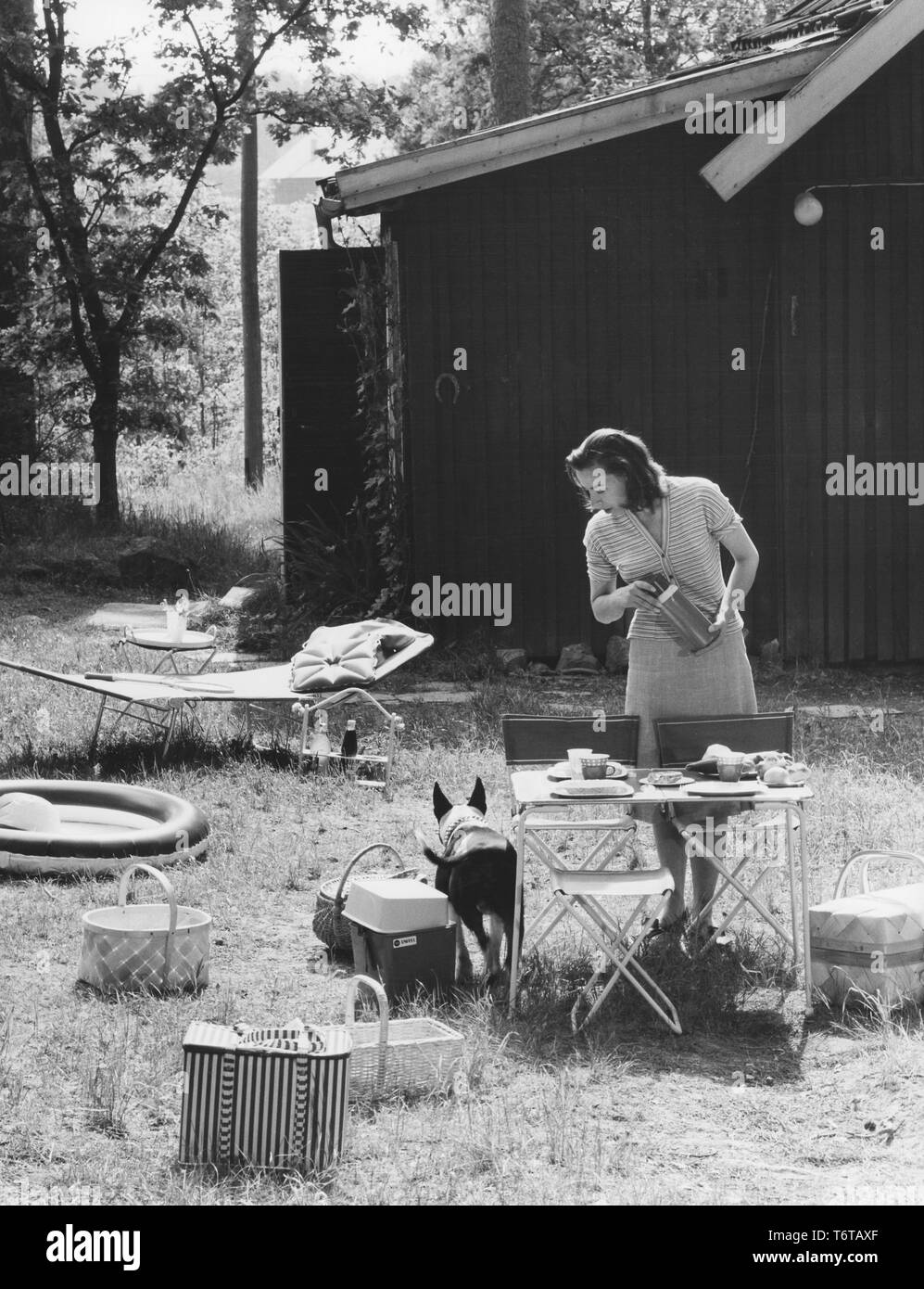 Camping in den 1960er Jahren. Beim Camping in den 1960er Jahren Plastikbecher und andere Campingausrüstung waren manchmal verpackt ist und in besonderen Fällen transportiert. Eine praktische wie abgebildet und eine Frau für eine Mahlzeit in der Natur. Die Stühle und der Tisch sind typische camping Falten, die leicht gespeichert und transportiert werden, nicht zu viel Raum in den Kofferraum des Autos. Jede sichtbare Objekt im Bild stellt die 1960er Dekade. Sowohl in Material und Design. Schweden 1963 Stockfoto