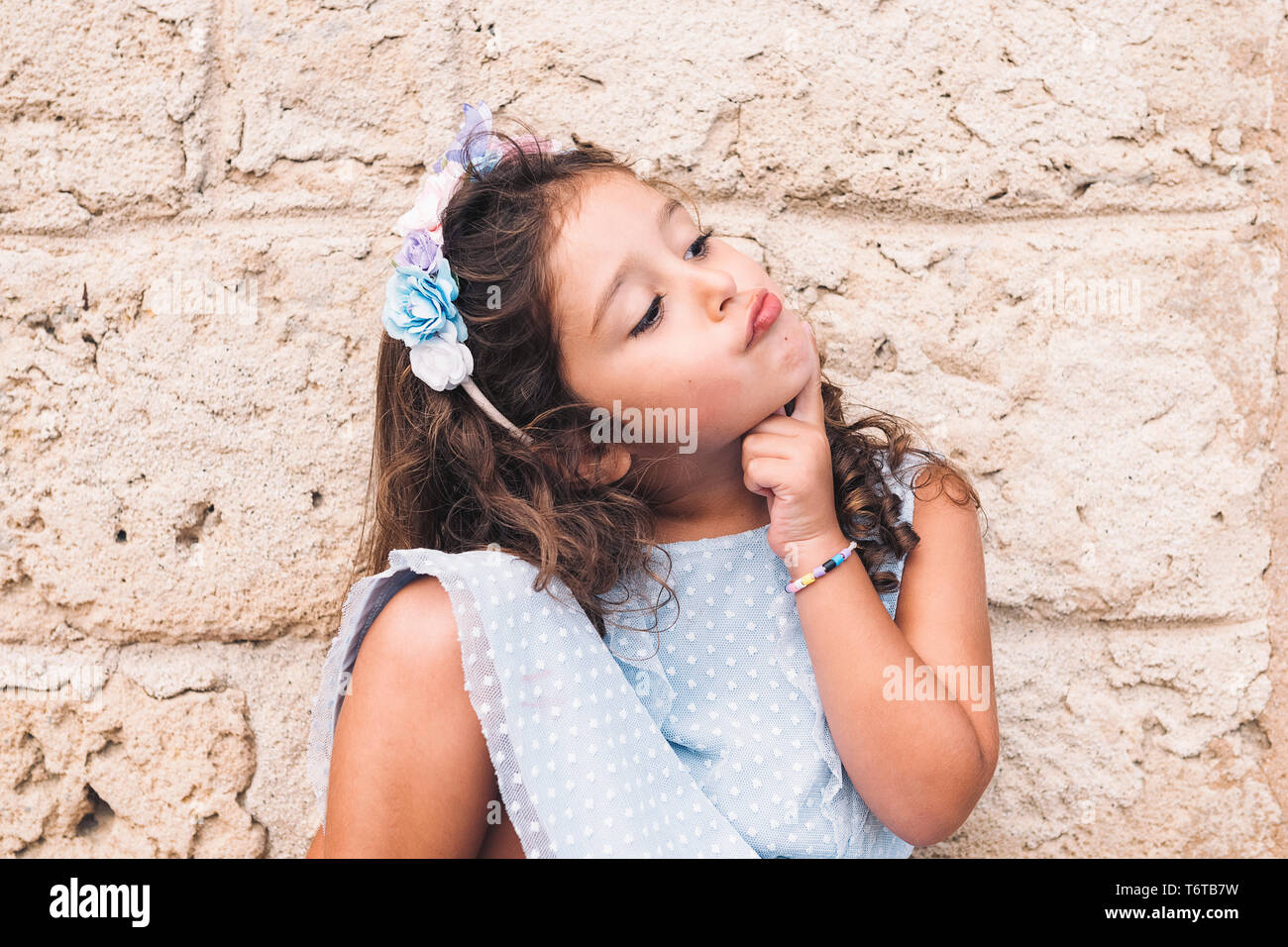 Lustige kleine Mädchen tun, wie sie denkt, ist vor einer Mauer aus Stein und trägt ein blaues Kleid und eine Blume Kopfbügel Stockfoto