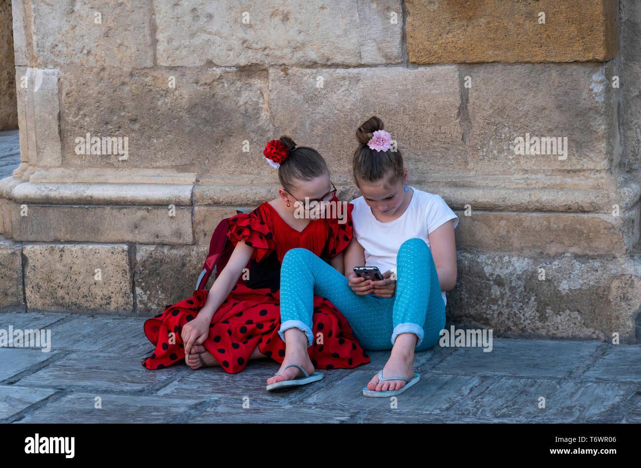 Zwei jugendliche Mädchen mit Blumen im Haar spielen mit einem Smartphone. Ein Mädchen trägt ein Flamenco Kleid Stockfoto