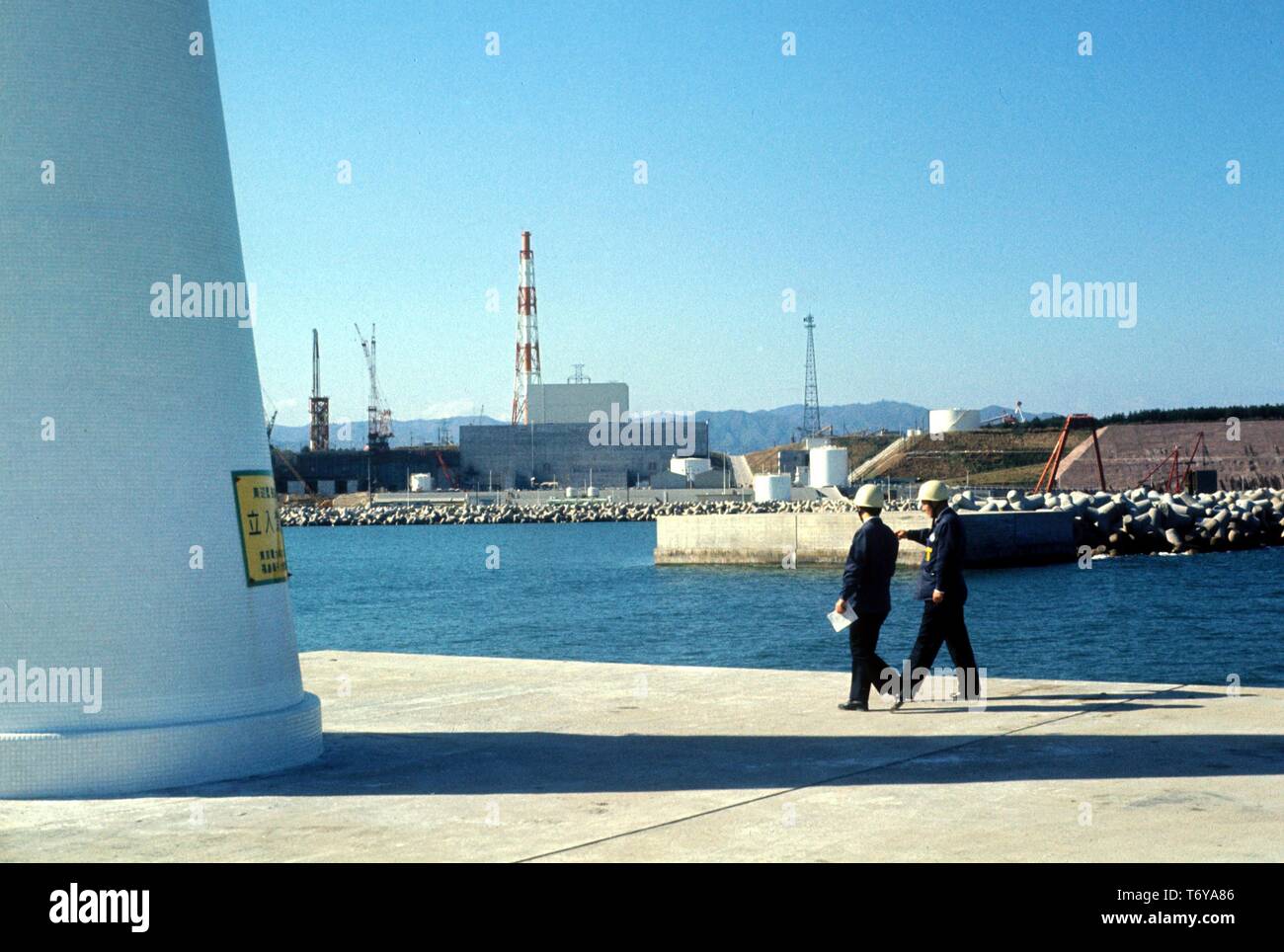 Zwei Männer tragen Bau Helme Spaziergang auf einem betonpfeiler, an einem sonnigen Tag, mit dem Kernkraftwerk Fukushima Daiichi im Hintergrund, Fukushima, Japan, 1970. Mit freundlicher Genehmigung des US-Ministeriums für Energie. () Stockfoto