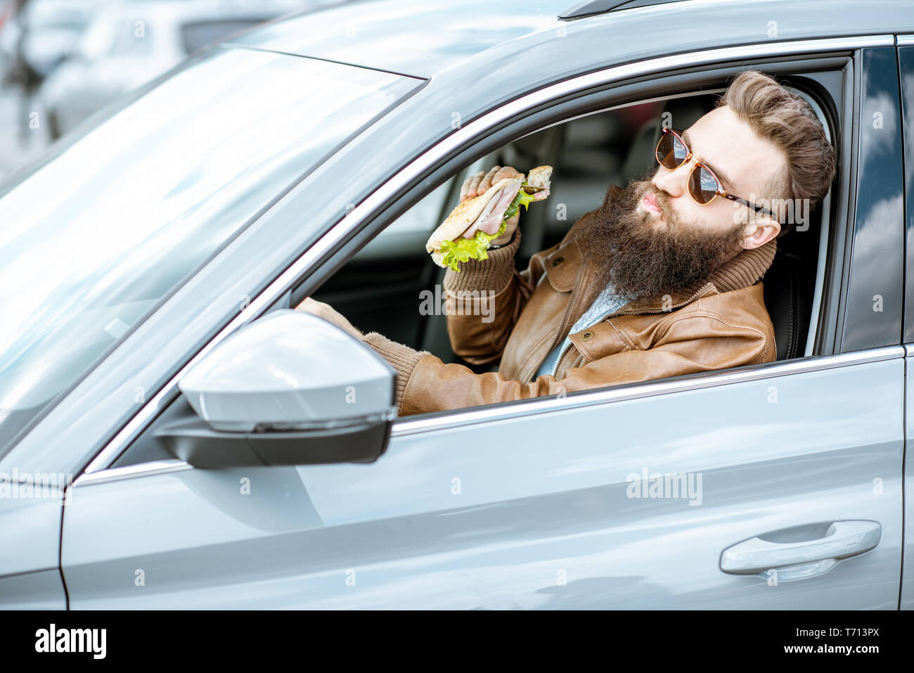 Stilvolle bärtiger Mann einen Snack mit leckeren Sandwich beim Autofahren in der Stadt Stockfoto