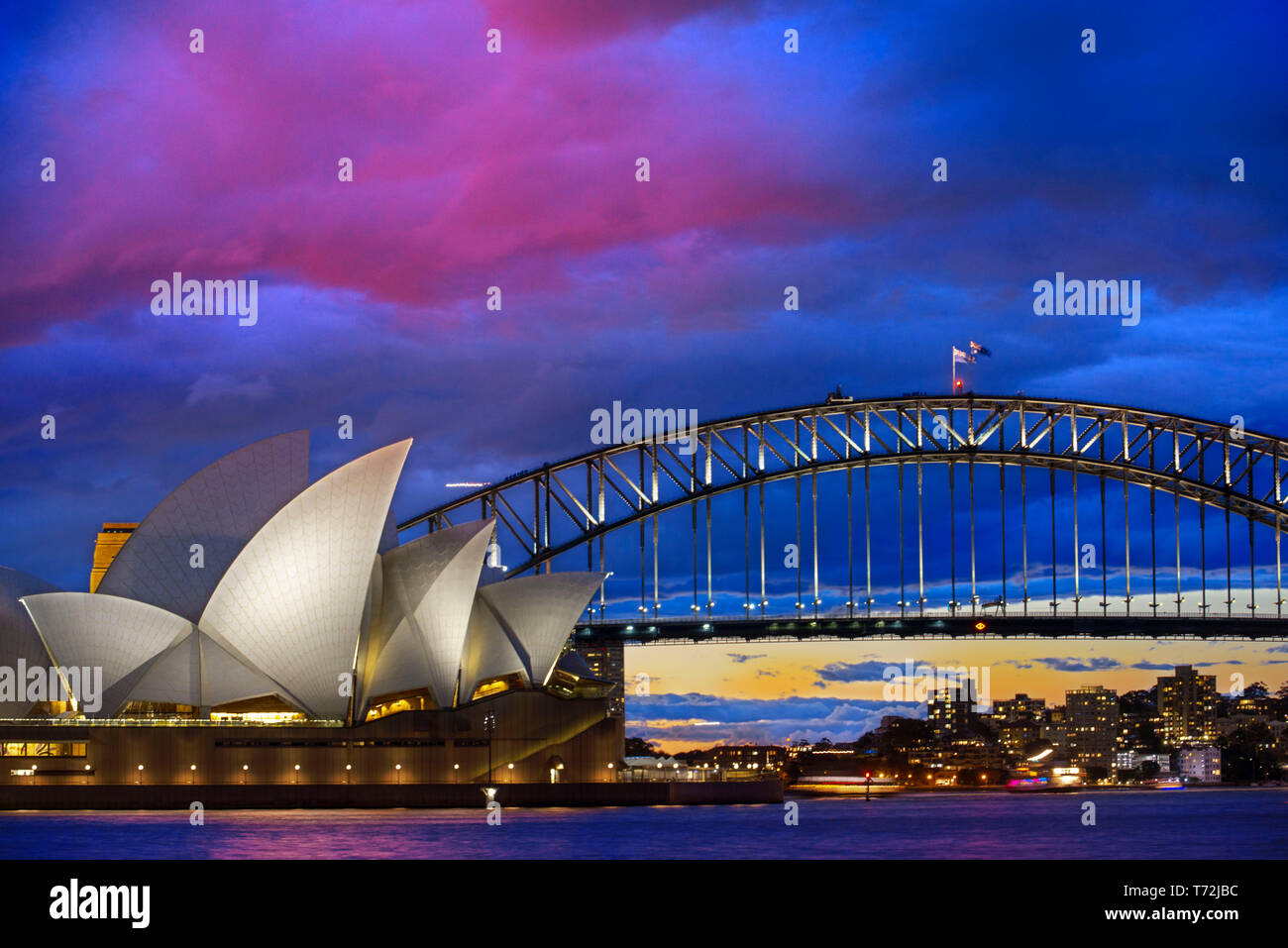 Welt berühmten Sydney Opera House und die Harbour Bridge bei Sonnenuntergang. Unscharfe Wolken und Lichter von Wahrzeichen spiegeln sich in fliessend Wasser des Hafens. Sydney, New Stockfoto