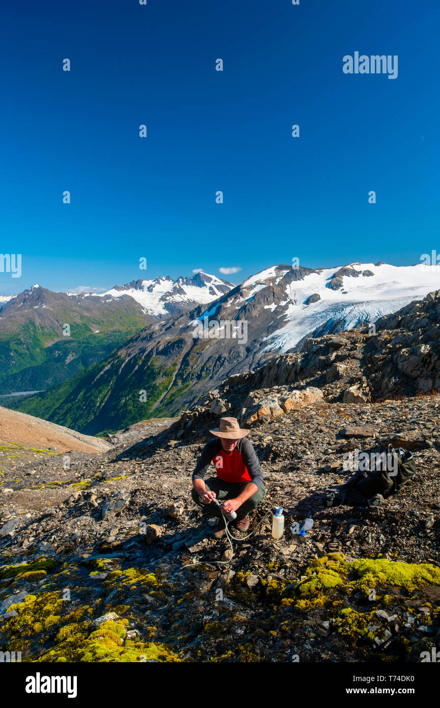 Ein Mann wird gesehen, Filtern von Wasser beim Wandern die herrliche Harding Icefield Trail im Kenai Fjords National Park mit einem hängenden Gletscher im Hintergrund Stockfoto