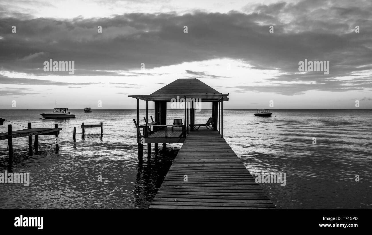 Schwarz-weiß-Bild eines Dock mit Sitzgelegenheiten und Boote Liegeplatz auf der ruhigen Wasser bei Sonnenuntergang; Roatan, Bay Islands, Honduras Stockfoto