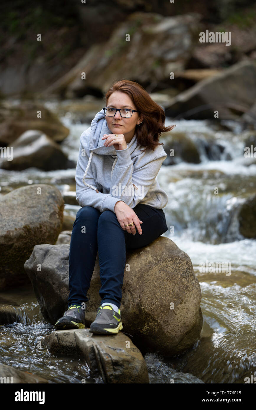Junge Frau sitzt auf einem Felsen in der Mitte von einem Fluss, während Sie Ihr Kinn mit einer Hand Stockfoto