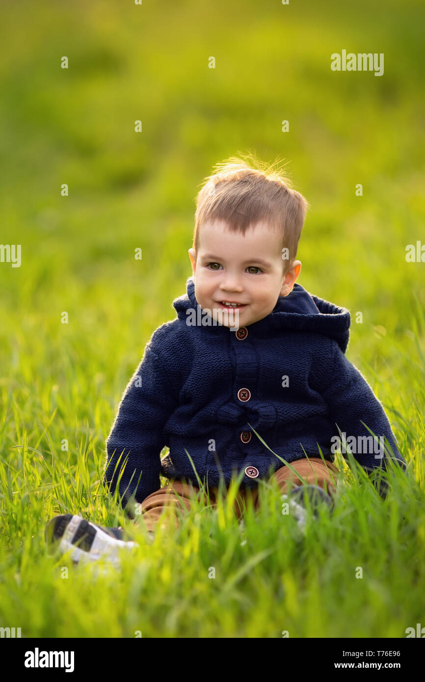 Lächelnd wenig Junge sitzt im grünen Gras an einem Frühlingstag. Schönes Kind Portrait im Freien Stockfoto