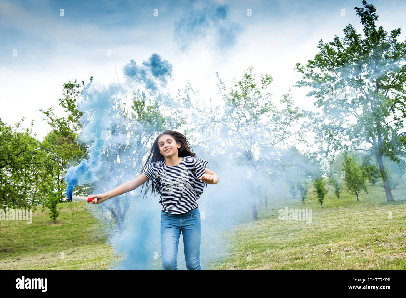 Junge Mädchen spielen mit einem blauen Rauch flare über das Gras in einem bewaldeten Park mit einem glücklichen Lächeln in die Kamera Stockfoto