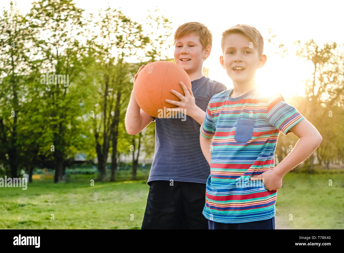 Zwei glückliche junge Freunde oder Brüder, stehend auf einem ländlichen Sportplatz Halten eines Basketball mit glücklichen Grinsen Hintergrundbeleuchtung durch das Licht der untergehenden s Stockfoto