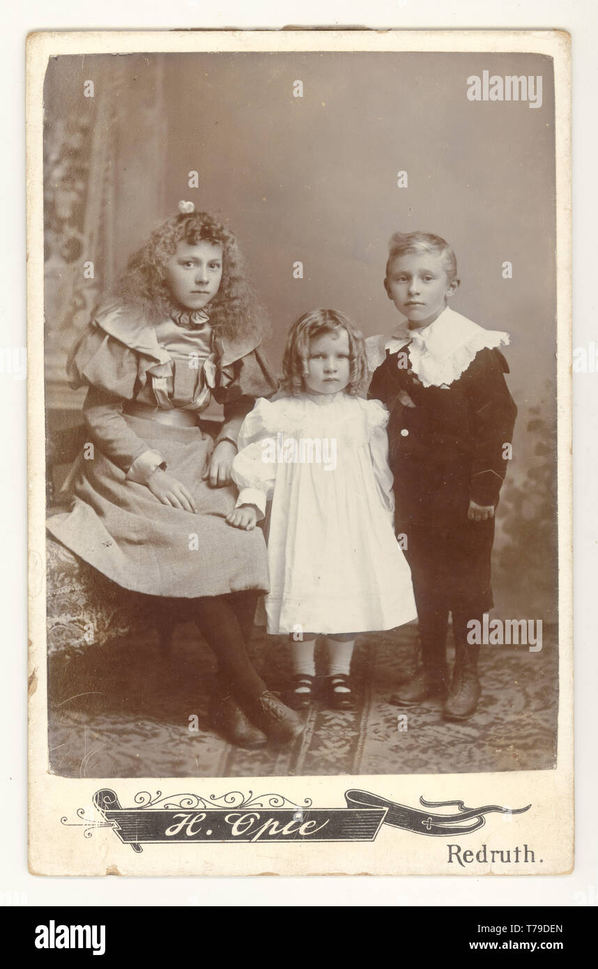 Originale viktorianische Kabinettkarte von 3 gut gekleideten, eleganten kornischen Kindern, Bruder mit Schwestern in hübschen Kleidern, aus den Studios von H. Opie, Redruth, Cornwall, Großbritannien um 1895,1896 Stockfoto