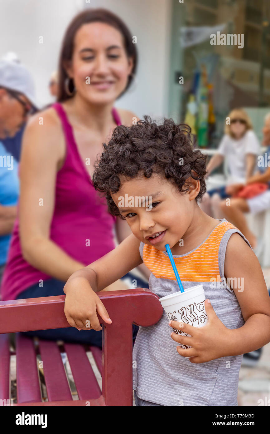 Ein Junge mit dem lockigen Haar grinst in die Kamera. ​A Toddler steht am Rand der Sitzbank mit Blick auf die Kamera, während Mama lächelt im Hintergrund. Stockfoto