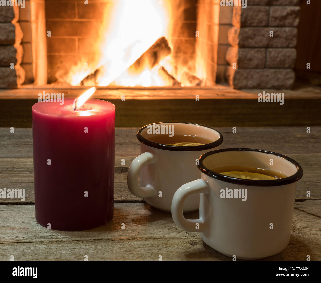 Weißer Becher mit heißem Tee und Kerzenschein in der Nähe von gemütlichem Kamin, im Landhaus, Winter Urlaub. Stockfoto