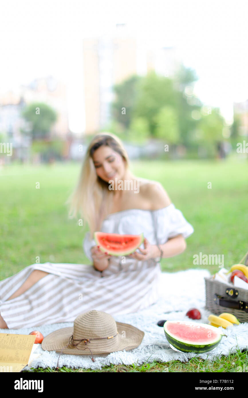 Junge Frau sitzt auf der Plaid in der Nähe von Früchten und Hut, Essen, Wassermelone, Gras im Hintergrund. Stockfoto
