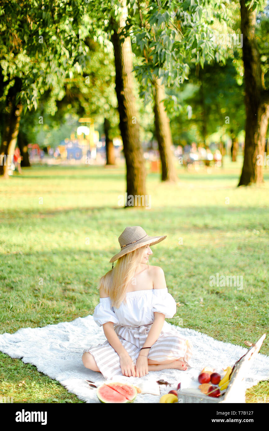 Junge Frau in aucasian Hut sitzt im Park auf Plaid in der Nähe von Obst, Gras im Hintergrund. Stockfoto