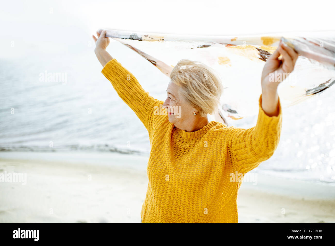 Ältere Frau im hellen Pullover genießen, Sea Breeze, holding Schal über dem Kopf am Sandstrand Stockfoto