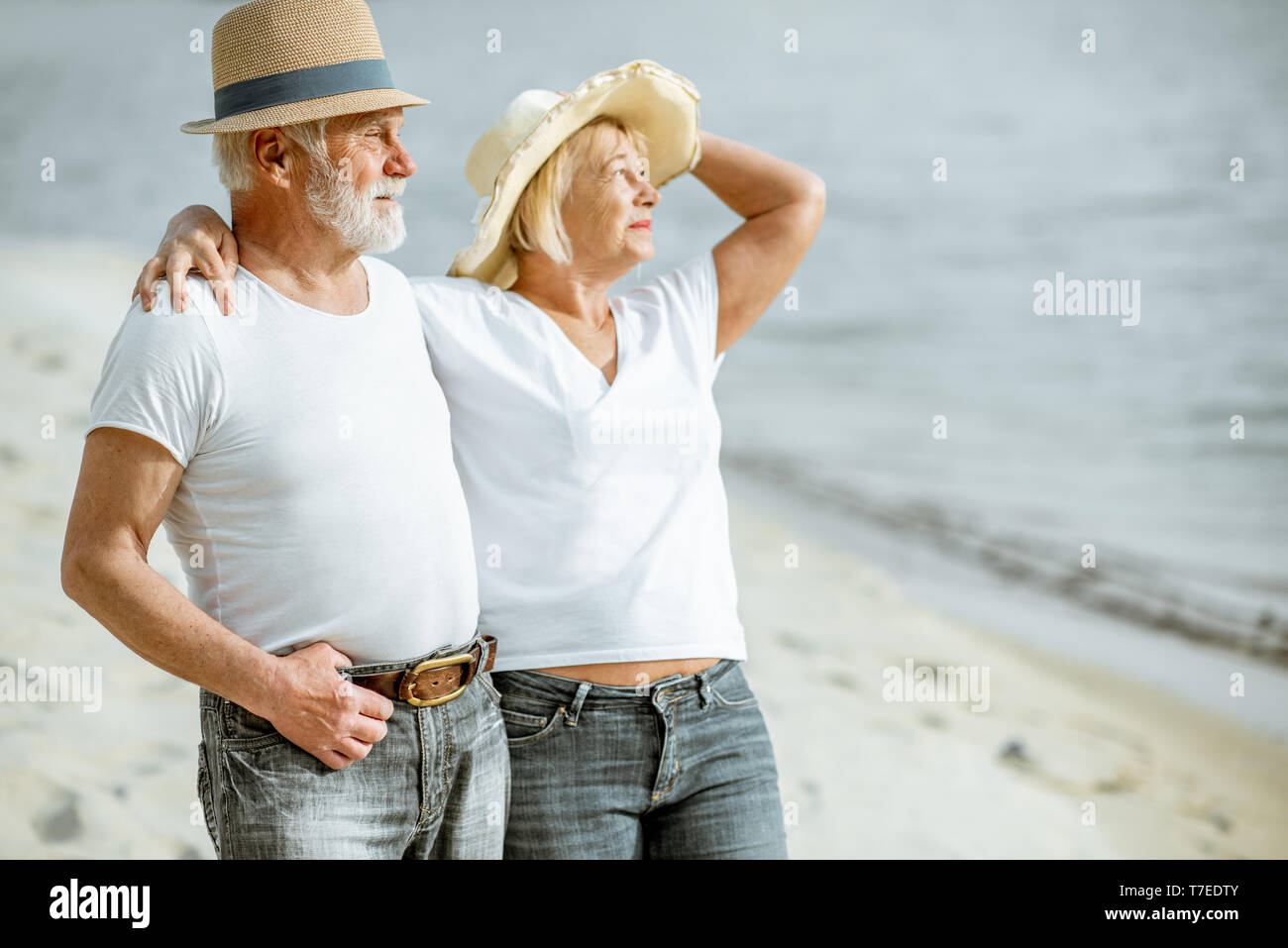 Gerne älteres Paar gekleidet in den weißen T-Shirts und Mützen zusammen zu Fuß am Sandstrand im Ruhestand Stockfoto