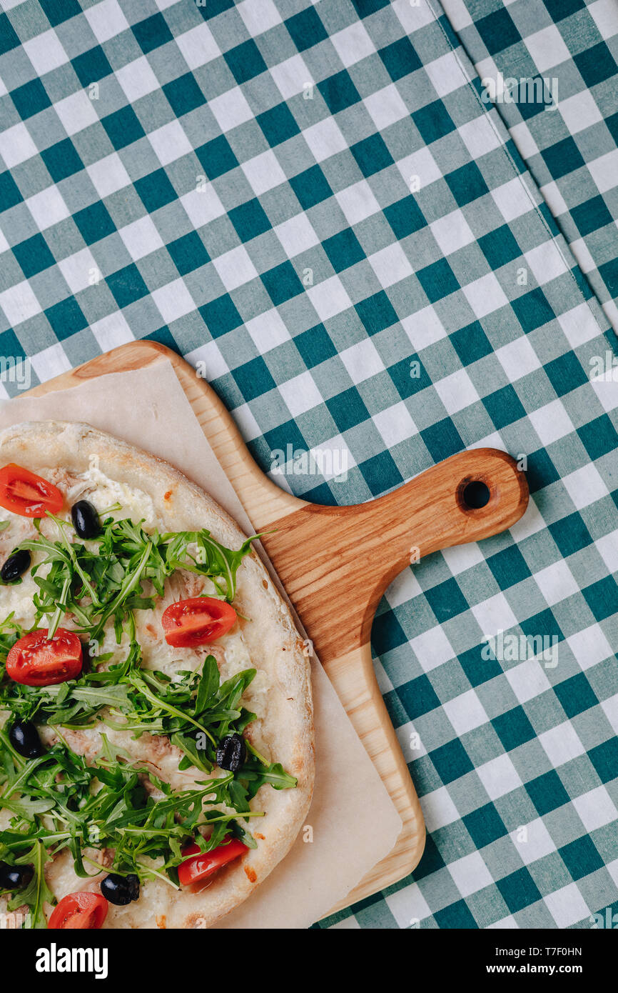 Neapolitanische Pizza auf Sahnesoße mit Thunfisch, Käse, Rucola, Basilikum, Tomaten, Oliven, bestreut mit Käse auf einem Holztisch auf einer Tischdecke in einer Zelle Stockfoto