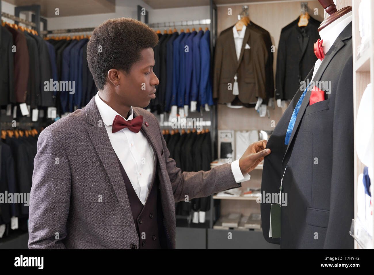 Junge afrikanische Mann in Boutique mit Kleidung für Männer in der Nähe von Mannequin, an der Jacke. Kunden Shop im weißen Hemd mit roter Schleife binden, schwarze, graue Jacke waiscoat Auswahl von eleganten Anzug. Stockfoto