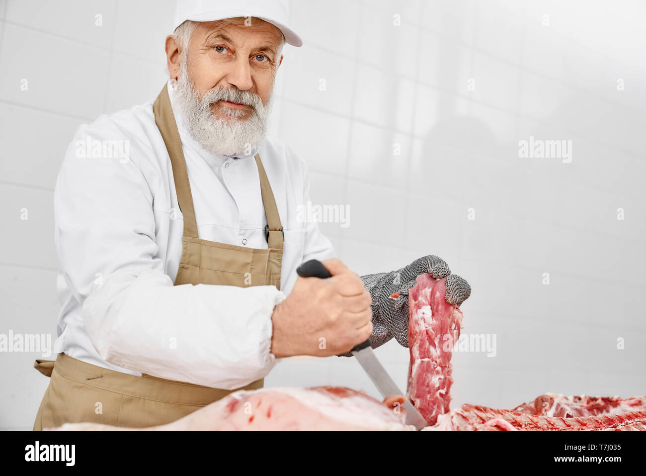 Gut aussehender älterer Mann arbeitet als Metzger holding Messer und Fleisch, an der Kamera schaut. Arbeitnehmer tragen in weiße Uniform, braune Schürze und spezielle Handschuhe schneiden frisches Schweinefleisch Schlachtkörper. Stockfoto