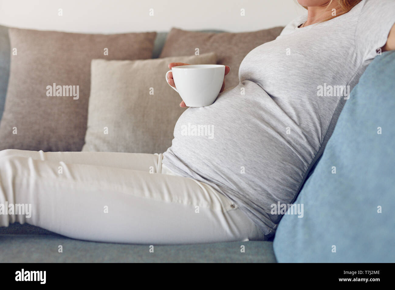 Schwangere Frau entspannt auf einem Sofa mit einer großen Tasse heißen Getränk in einer Nahaufnahme Seite Blick auf Ihr Baby bump und Hände halten die Schale Stockfoto