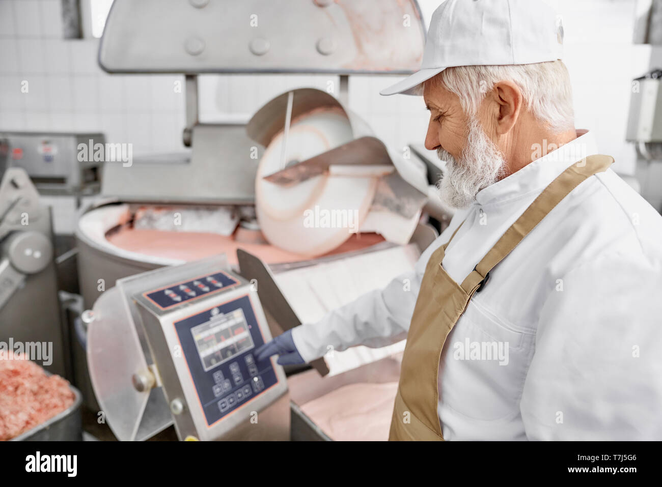 Älterer mann Arbeiter tragen in weißer Uniform, weiße Kappe, braun Schürze und Gummihandschuhe arbeiten an der Herstellung der Fleischerzeugung. Arbeiter in der Nähe von Ausrüstung für Schleifen Fleisch. Stockfoto