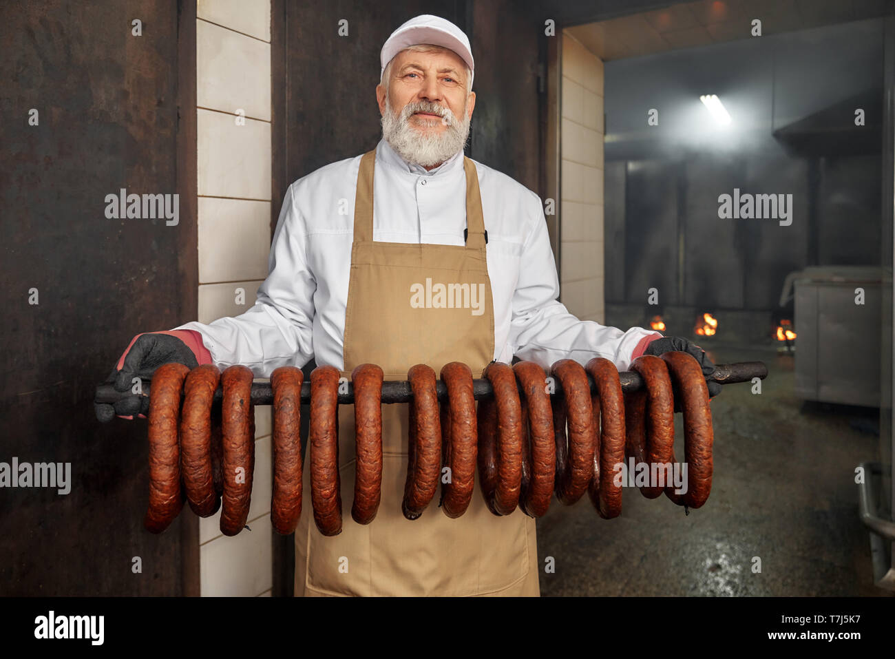 Der wurstherstellung. Ältere, gut aussehende Arbeiter in der räucherei mit geräucherter Wurst, an der Kamera schaut. Geräucherter sassages in Reihe hängen. Mann in weißer Uniform, braune Schürze, posieren. Stockfoto