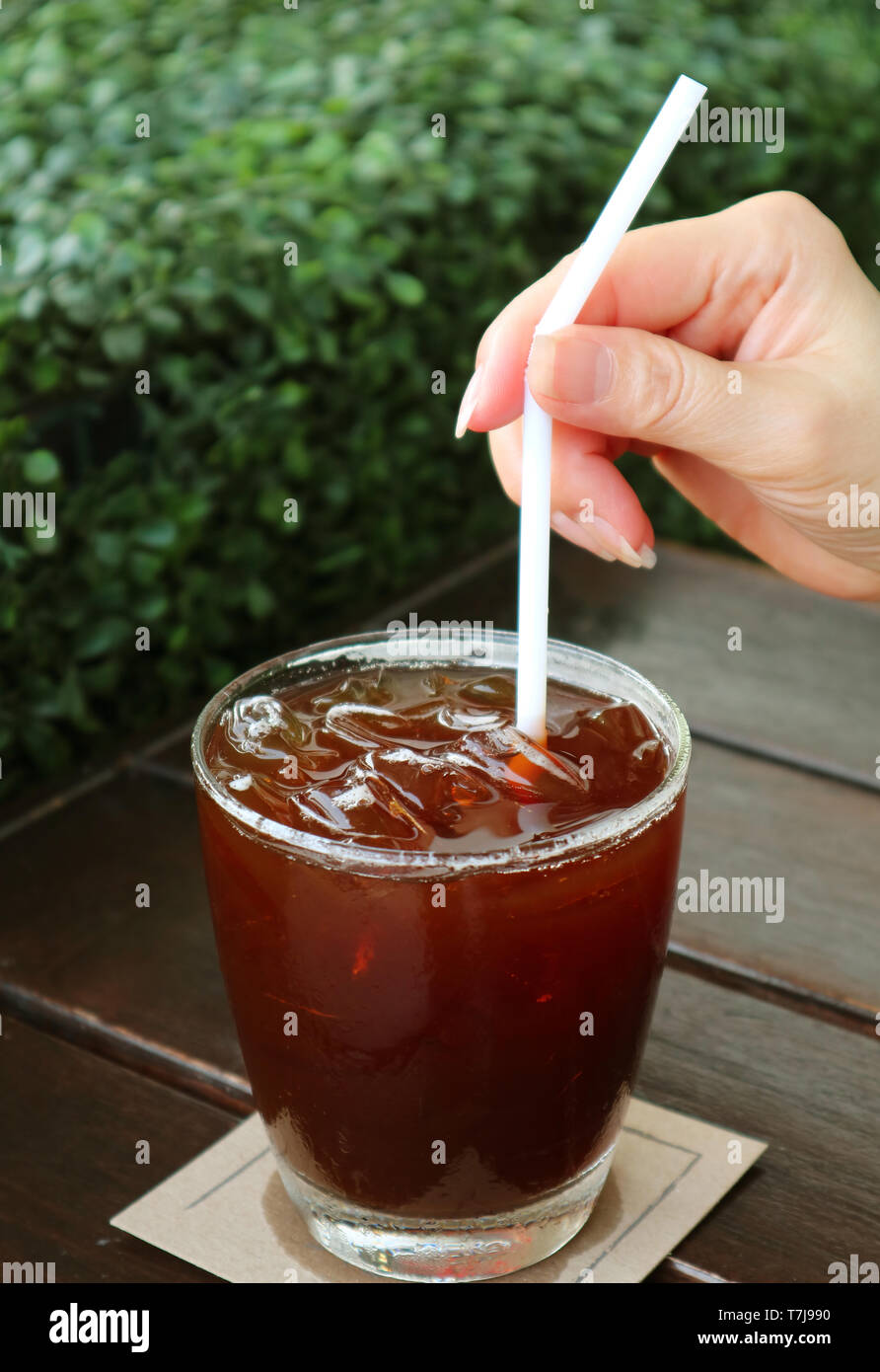 Woman's Hand mit einem Strohhalm in Eiskaffee Glas mit Blurry Green Bush im Hintergrund Stockfoto
