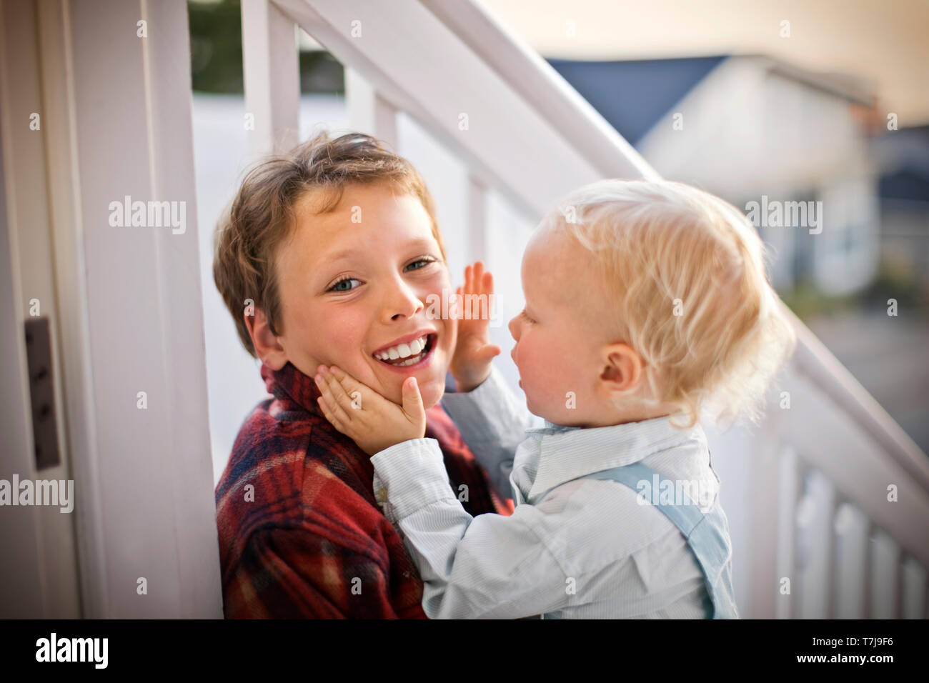 Porträt eines lächelnden Jungen sein Gesicht von seinem jüngeren Bruder berührt, während auf der Veranda seines Hauses sitzen. Stockfoto