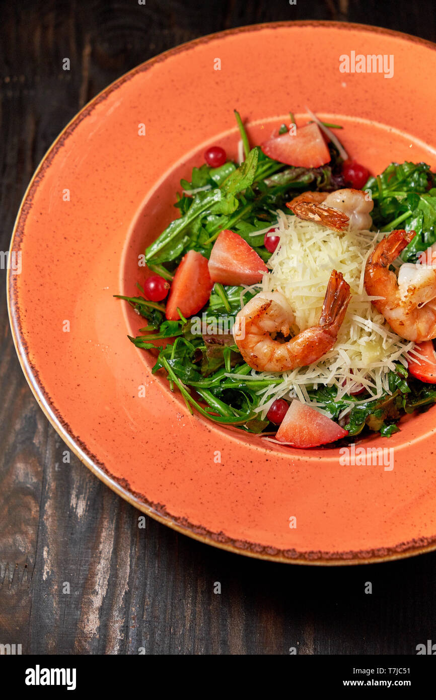 Frischer Salat mit Ruccola, Garnelen und Erdbeeren auf einem orangefarbenen Platte. Dunklen Hintergrund. Restaurant Menü Stockfoto