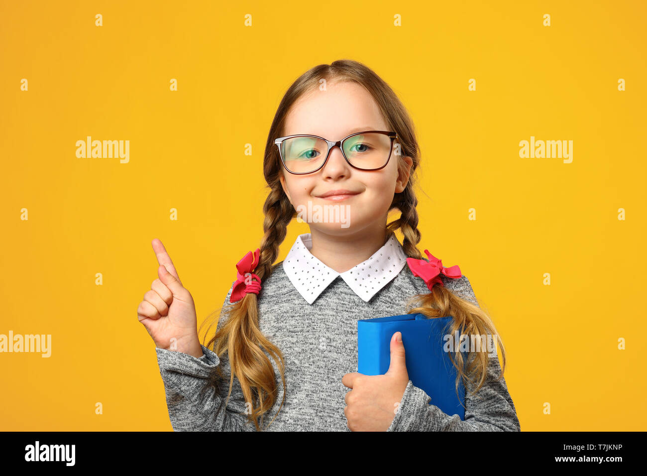 Closeup Portrait kleines Mädchen Schulmädchen auf gelben Hintergrund. Ein Kind von der Grundschule hält ein Lehrbuch und zeigt mit dem Finger nach oben. Das Konzept der Ed Stockfoto