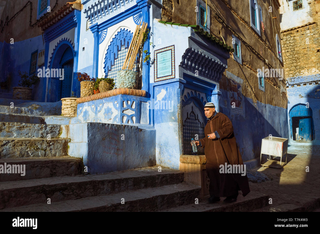 Chefchaouen, Marokko: ein Mann mit einem djellaba füllt eine Flasche Wasser aus einem Brunnen in der blau getünchten Gassen der Medina, der Altstadt. Stockfoto