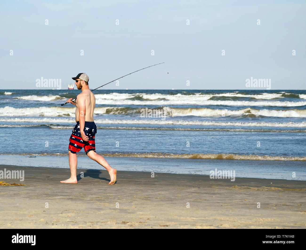 Ein Fischer mit Köder baumeln von der Pole in Shorts und Ball cap Spaziergänge am Strand neben dem Wasser in Port Aransas, Texas USA. Stockfoto