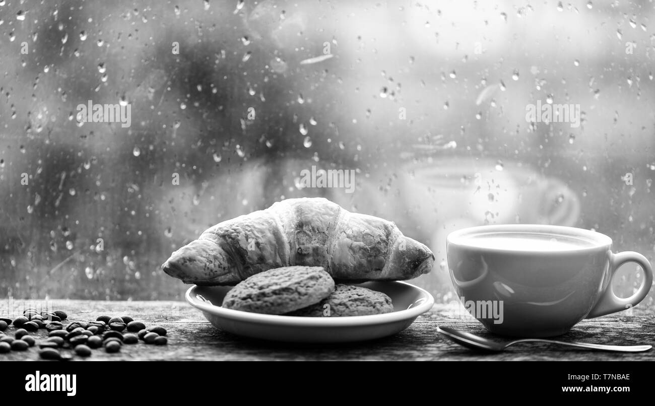 Genießen Kaffee an regnerischen Tag. Kaffee Zeit an regnerischen Tag. Frisch gebrühter Kaffee in weiße Tasse oder Becher auf der Fensterbank. Nasses Glas Fenster und heiße Tasse Koffein trinken. Trinken Kaffee mit Croissant Dessert. Stockfoto