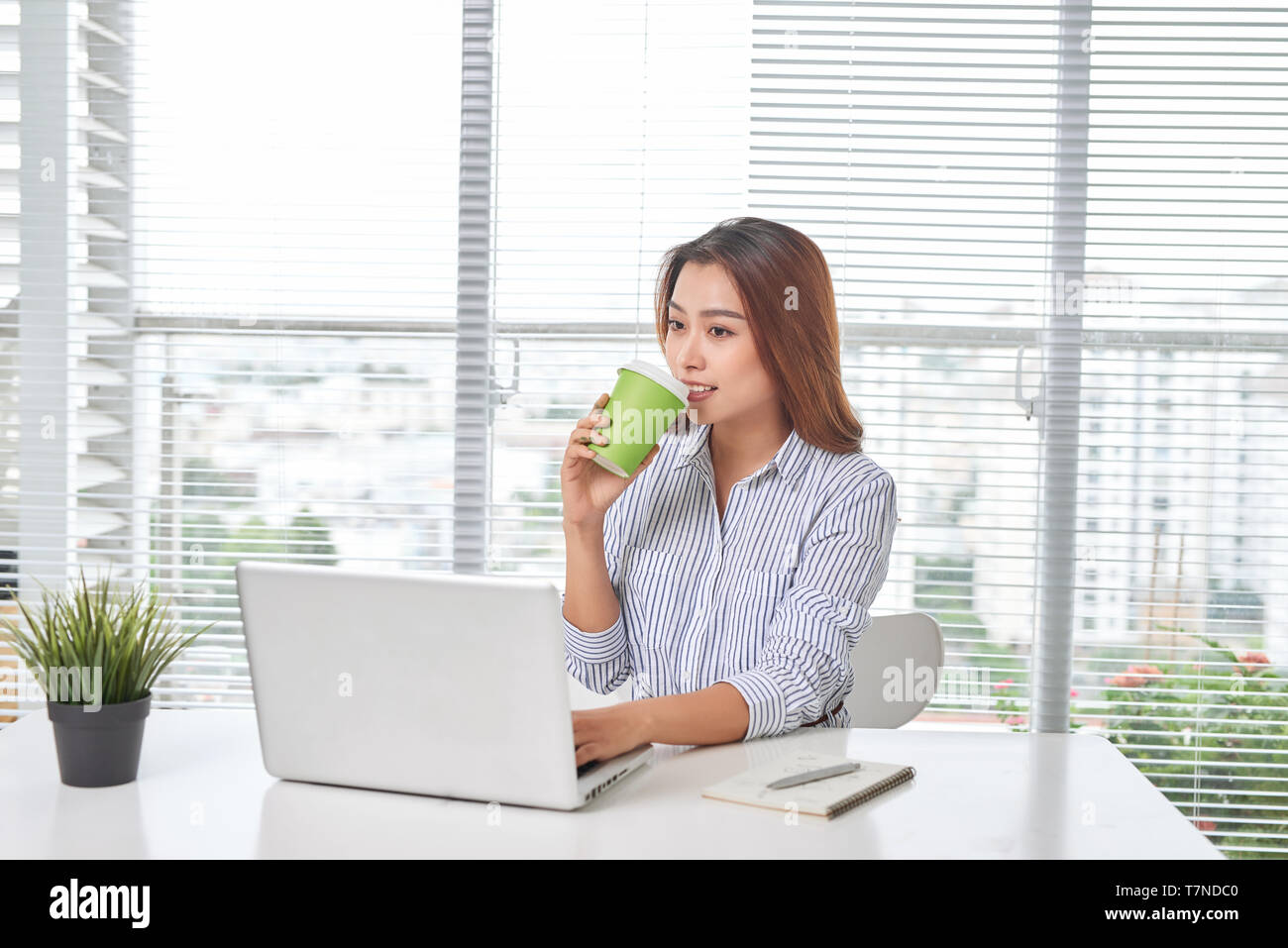 Schöne junge Frau am Tisch sitzend, im Home Office holding Tasse Kaffee, mit Laptop. Stockfoto