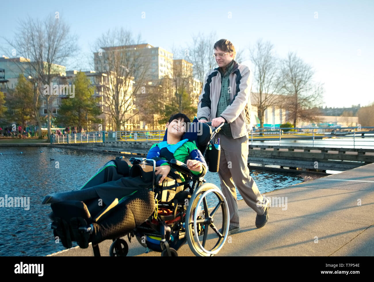 Kaukasische Vater wandern Behinderte biracial jungen Sohn im Park von lakle draußen im frühen Frühjahr oder Sommer. Kind hat zerebrale Lähmung. Stockfoto