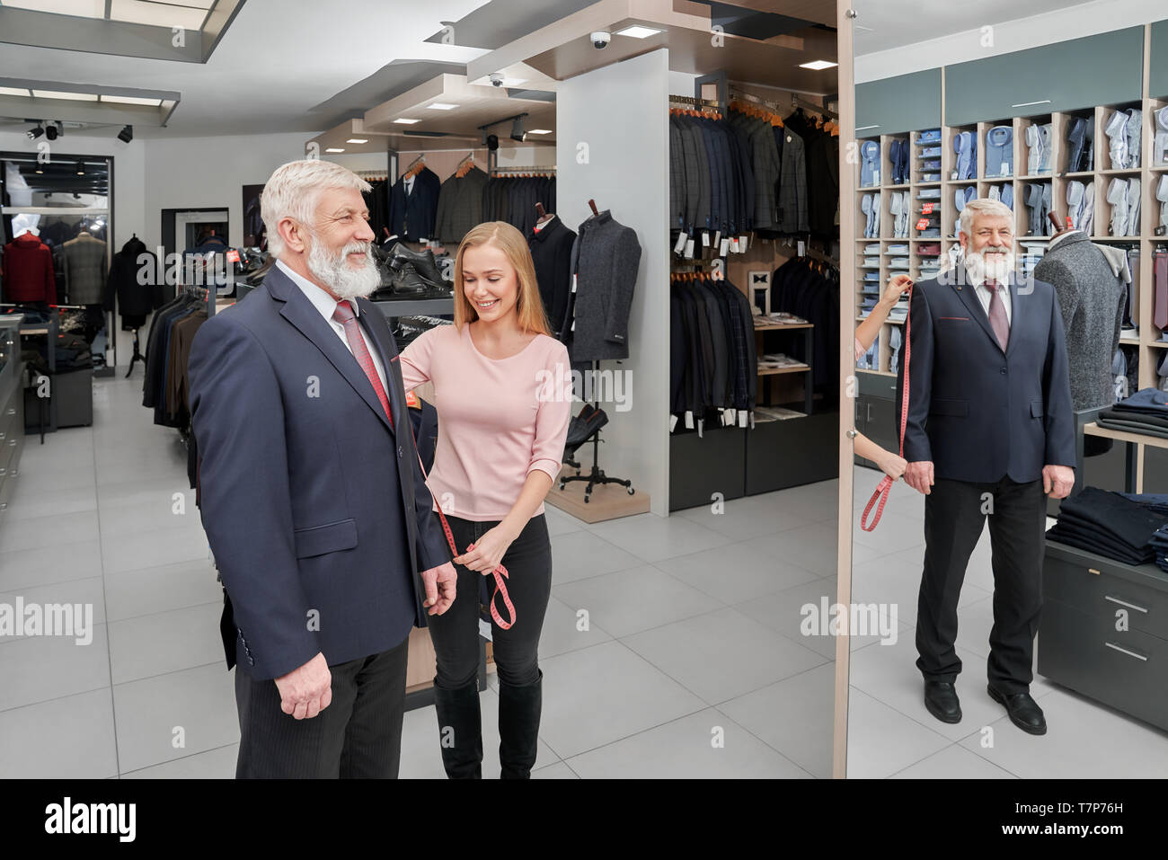 Seitenansicht des Großvaters tragen smart Klage auf Spiegel und lächelt, während die weiblichen assistant Meßhülse. Stilvolle männlichen Klienten die Auswahl und Kauf neuen Outfit. Konzept der Verschraubung und Kleidung. Stockfoto