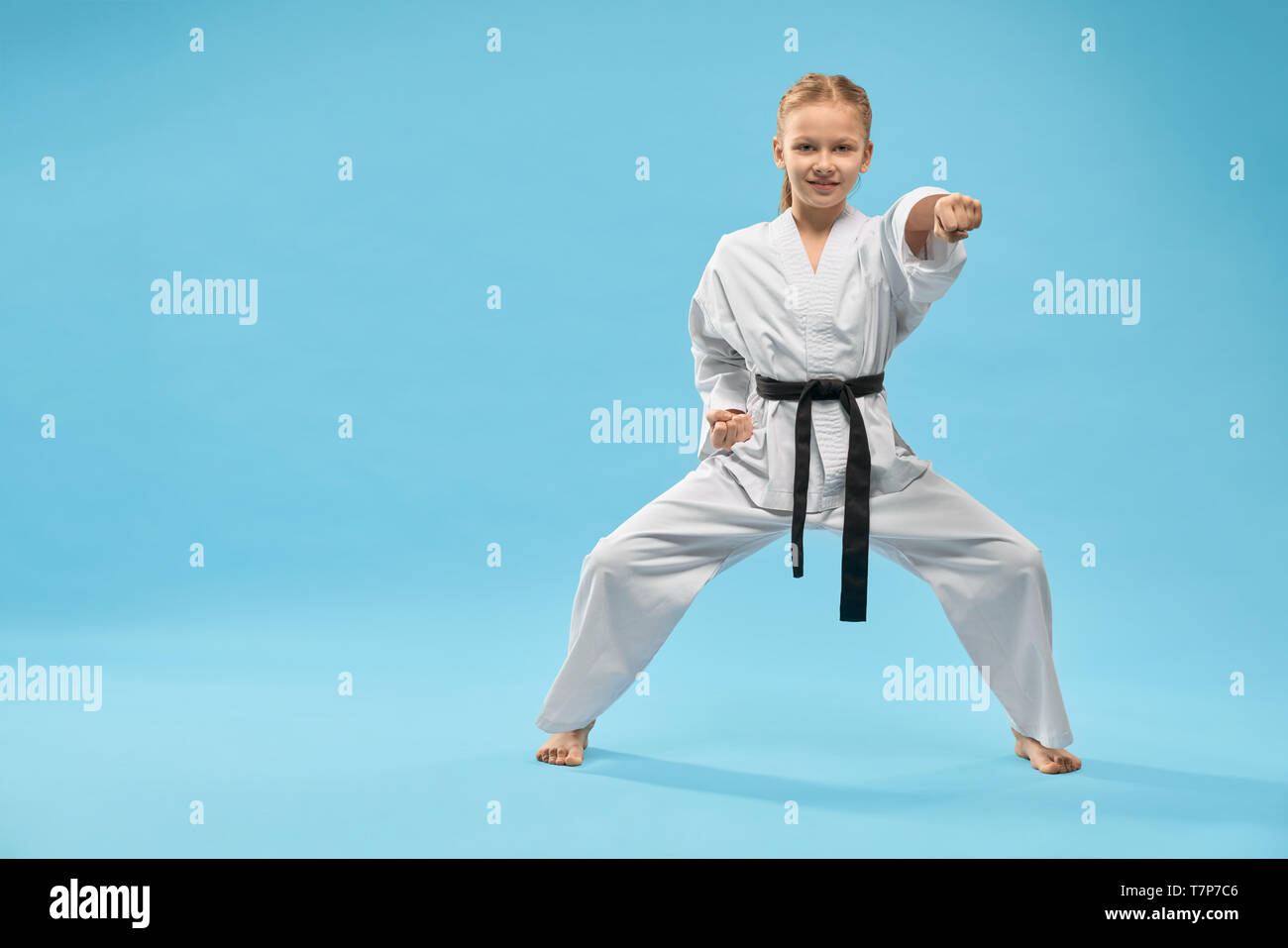 Vorderansicht der weiblichen Teenager stehend an Haltung und Stanzen auf blauem Hintergrund isoliert. Lächelnde Mädchen an Kamera und posiert im Studio. Kind kämpfen und Ausübung von Karate. Konzept der Sport. Stockfoto