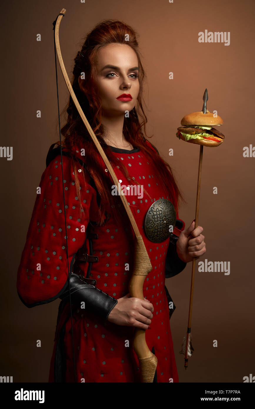 Portrait von bestimmt Kämpferin mit roter Rüstung, Pfeil und Bogen mit Hamburger im Studio. Spartanischen Krieger beiseite und auf isolierten Hintergrund posiert. Konzept der Macht und Essen. Stockfoto