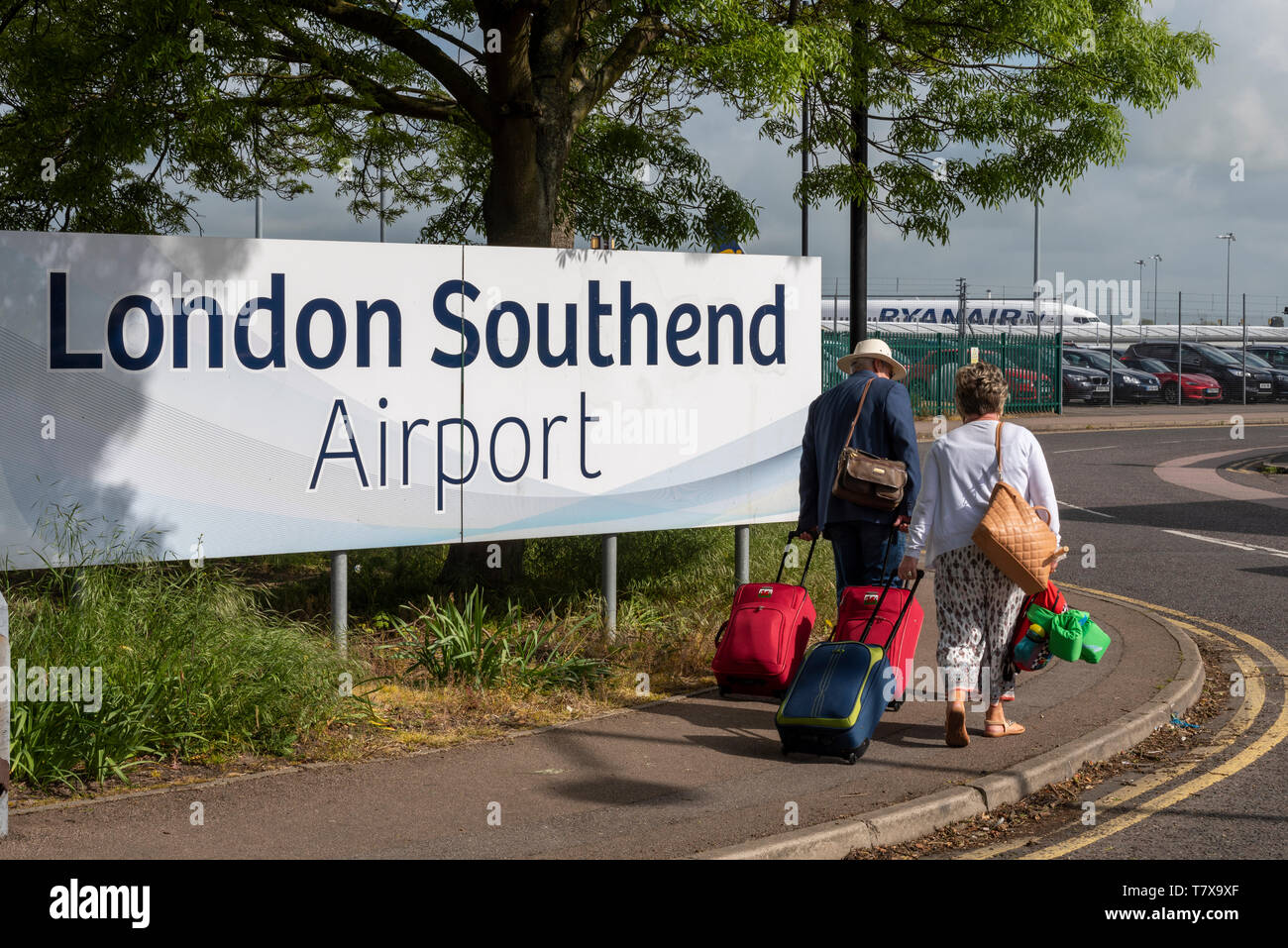Familie von Passagieren, die am Einfahrtsschild des London Southend Airport vorbeilaufen, mit dem Flugzeug von Ryanair dahinter. Southend on Sea, Essex, Großbritannien Stockfoto