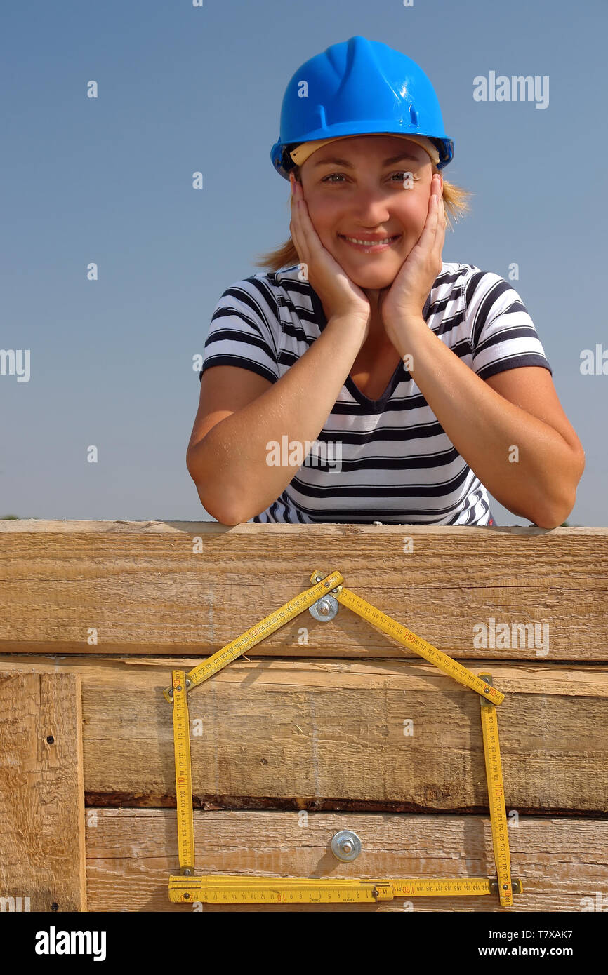 Junge weibliche trägt blaue Helm über gelbe Haus geformte Holz- Zollstock posing angebracht zu den hölzernen Verschalung Wand über blauen Himmel Stockfoto