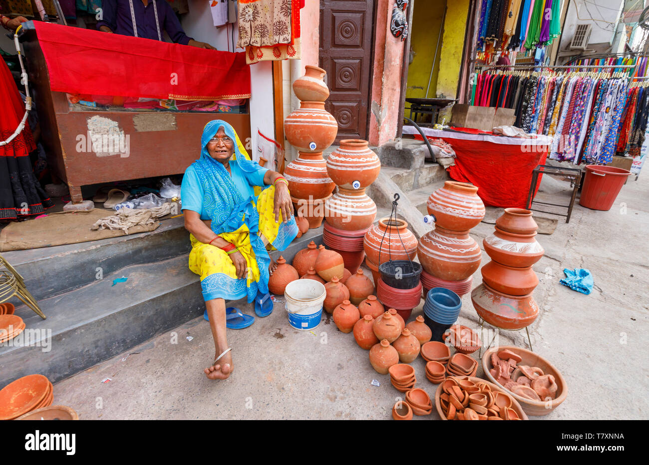 Street Scene, Mahipalpur district, New Delhi Suburb: Eine alte Frau sitzt auf dem Bürgersteig außerhalb Kleidung kaufen mit einem Display von keramischen Töpfen Stockfoto