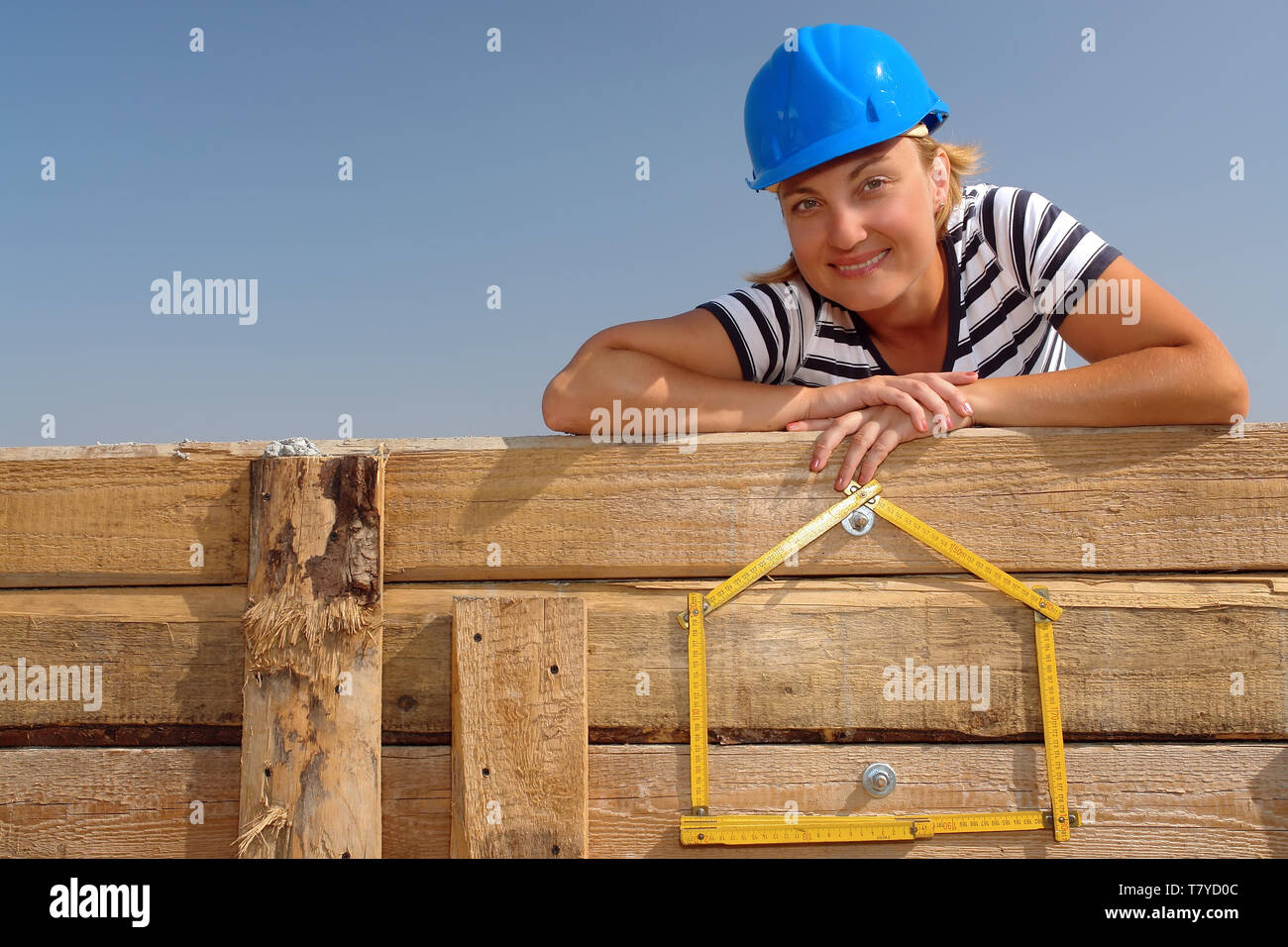Junge weibliche trägt blaue Helm über gelbe Haus geformte Holz- Zollstock posing angebracht zu den hölzernen Verschalung Wand über blauen Himmel Stockfoto