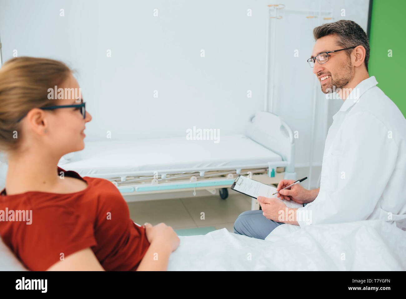 Männlicher Arzt in einem Gespräch mit einem lächelnden Patient in einem Krankenhaus Bett liegen Stockfoto