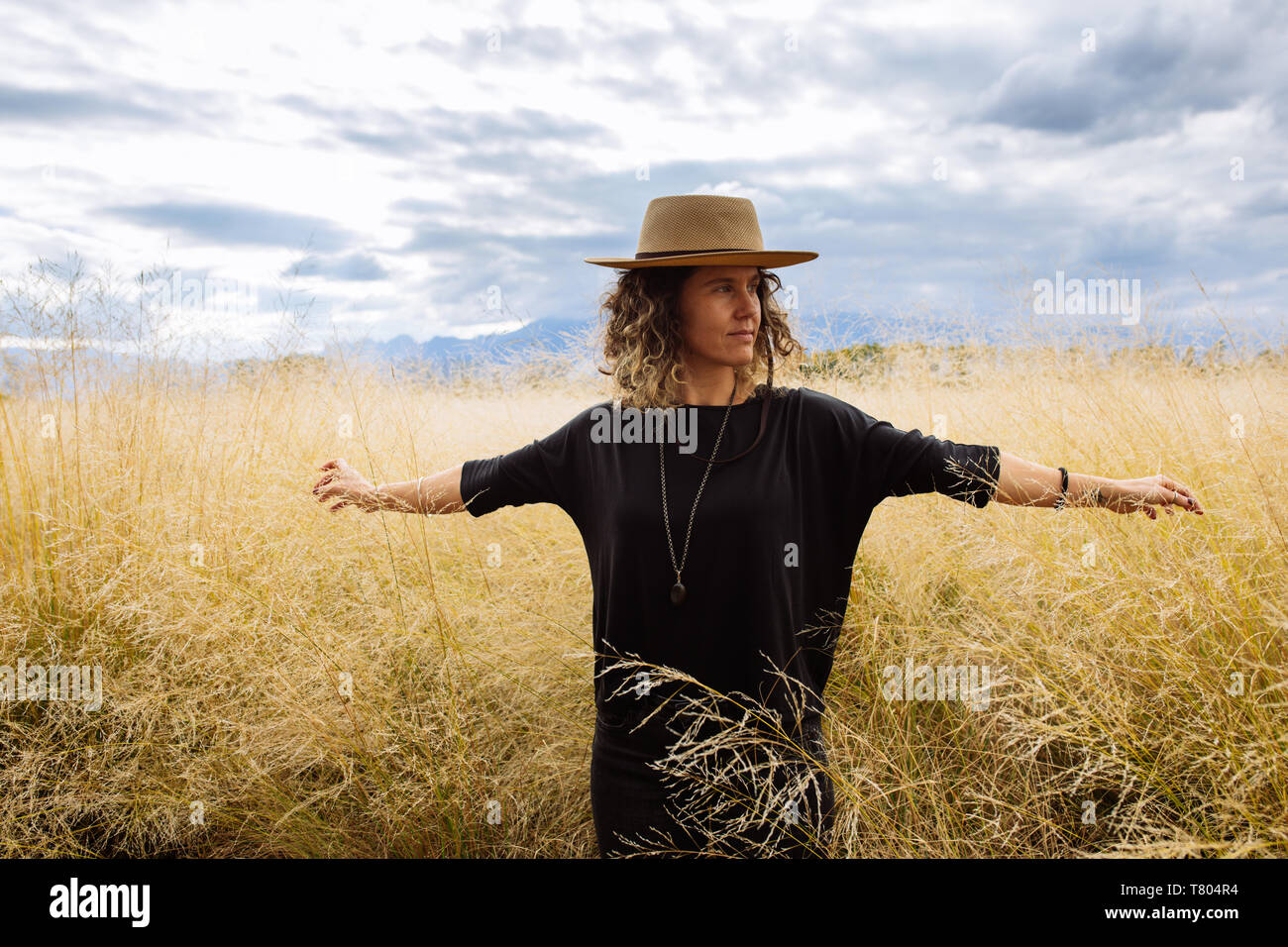 Frau, Mitte der 30er Jahre, die sich mit offenen Armen mit einem Hut in einem gelben Gras Feld in Bodega Septima Weingut in Mendoza, Argentinien Stockfoto